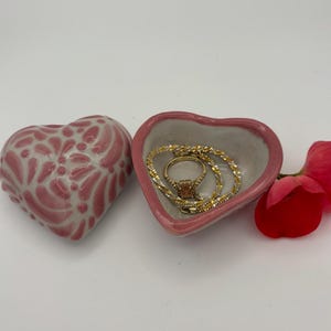 Pode incluir: Uma tigela de joias em forma de cora&ccedil;&atilde;o rosa com um anel e um colar de ouro dentro. Uma segunda caixa de bugigangas em forma de cora&ccedil;&atilde;o com um design floral branco e rosa est&aacute; ao lado do prato. Uma flor vermelha est&aacute; &agrave; direita.