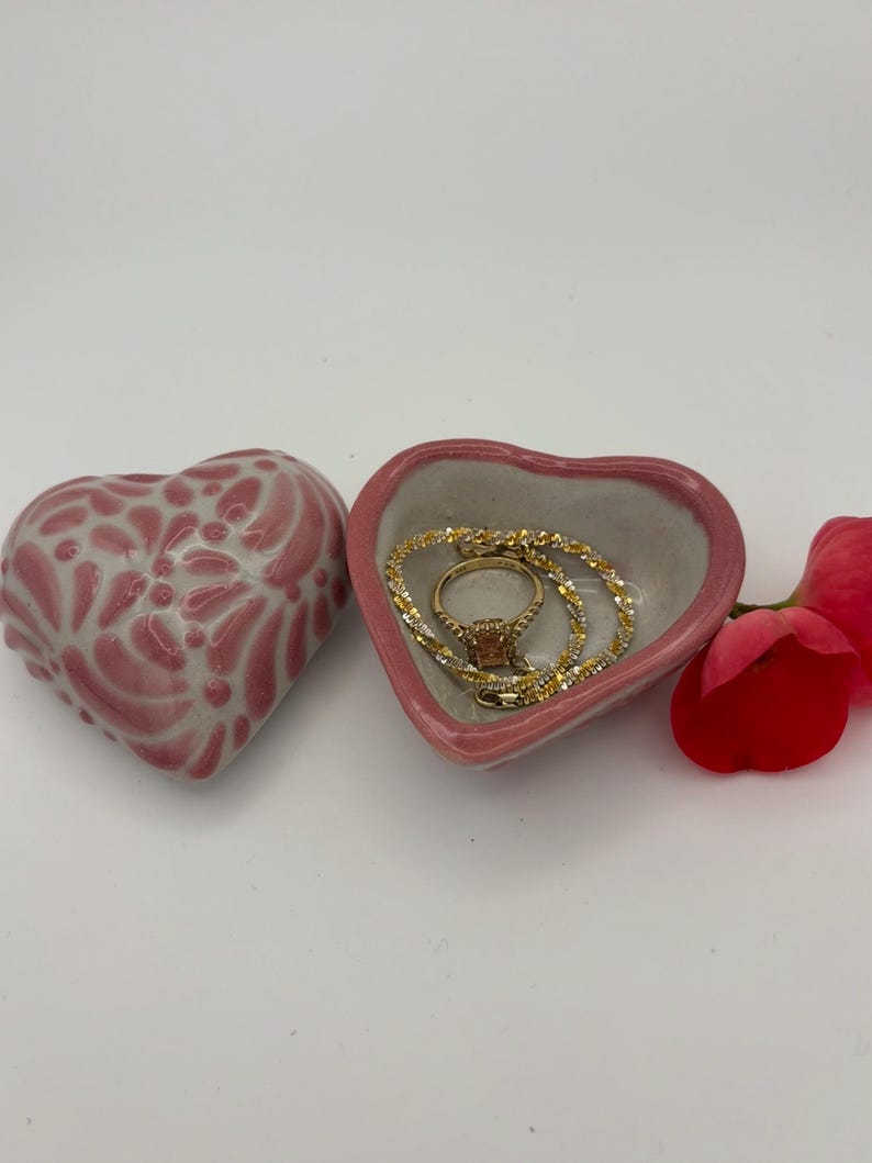 Pode incluir: Uma tigela de joias em forma de cora&ccedil;&atilde;o rosa com um anel e um colar de ouro dentro. Uma segunda caixa de bugigangas em forma de cora&ccedil;&atilde;o com um design floral branco e rosa est&aacute; ao lado do prato. Uma flor vermelha est&aacute; &agrave; direita.