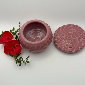 May include: A pink ceramic jar with a matching lid, both adorned with a floral pattern. The jar is open, revealing its interior. Red flowers and green leaves are placed near the jar.
