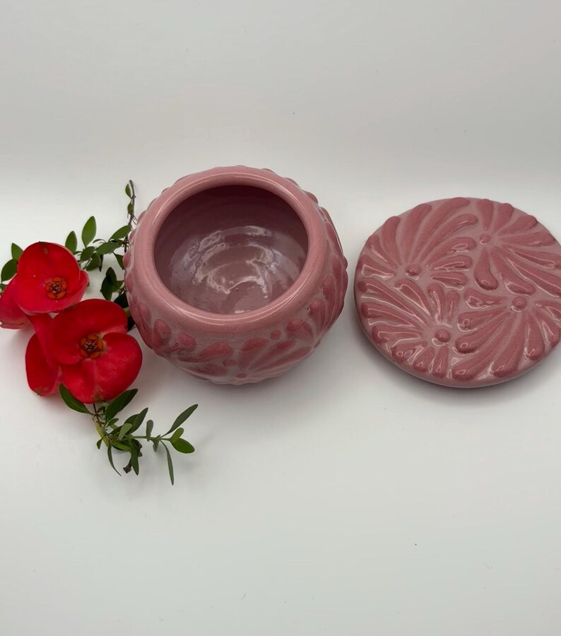 May include: A pink ceramic jar with a matching lid, both adorned with a floral pattern. The jar is open, revealing its interior. Red flowers and green leaves are placed near the jar.