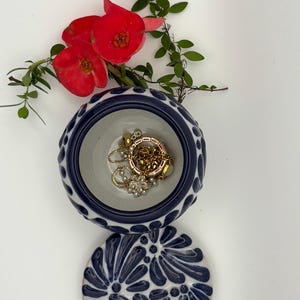 May include: A small, round, ceramic jewelry dish with a lid. The dish is white with a blue floral pattern and a blue rim. Inside the dish are gold necklaces and a flower. Red flowers and green leaves are arranged around the dish.