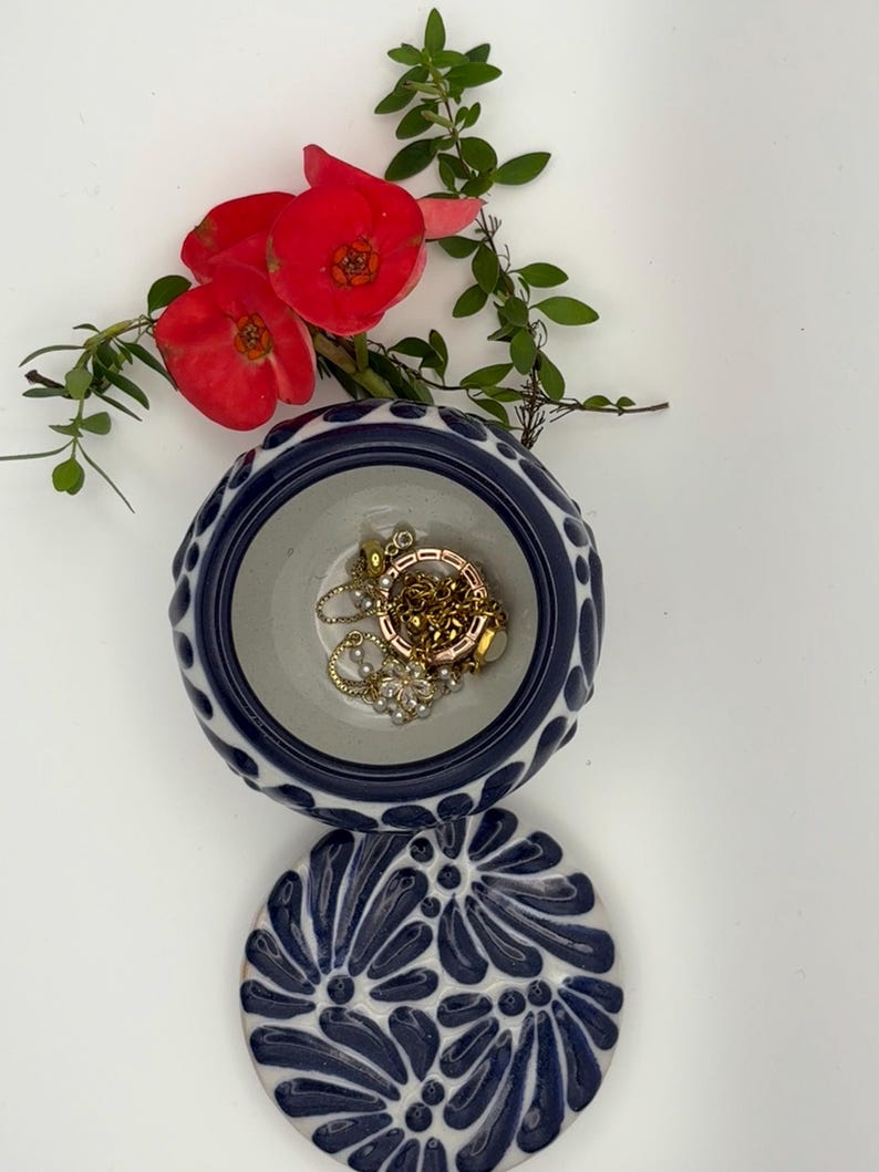 May include: A small, round, ceramic jewelry dish with a lid. The dish is white with a blue floral pattern and a blue rim. Inside the dish are gold necklaces and a flower. Red flowers and green leaves are arranged around the dish.