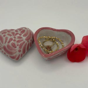 Pode incluir: Uma bandeja de cer&acirc;mica rosa em forma de cora&ccedil;&atilde;o com um anel e uma pulseira dourados dentro. Ao lado, uma segunda bandeja em forma de cora&ccedil;&atilde;o com um design floral. &Agrave; direita, uma rosa vermelha.