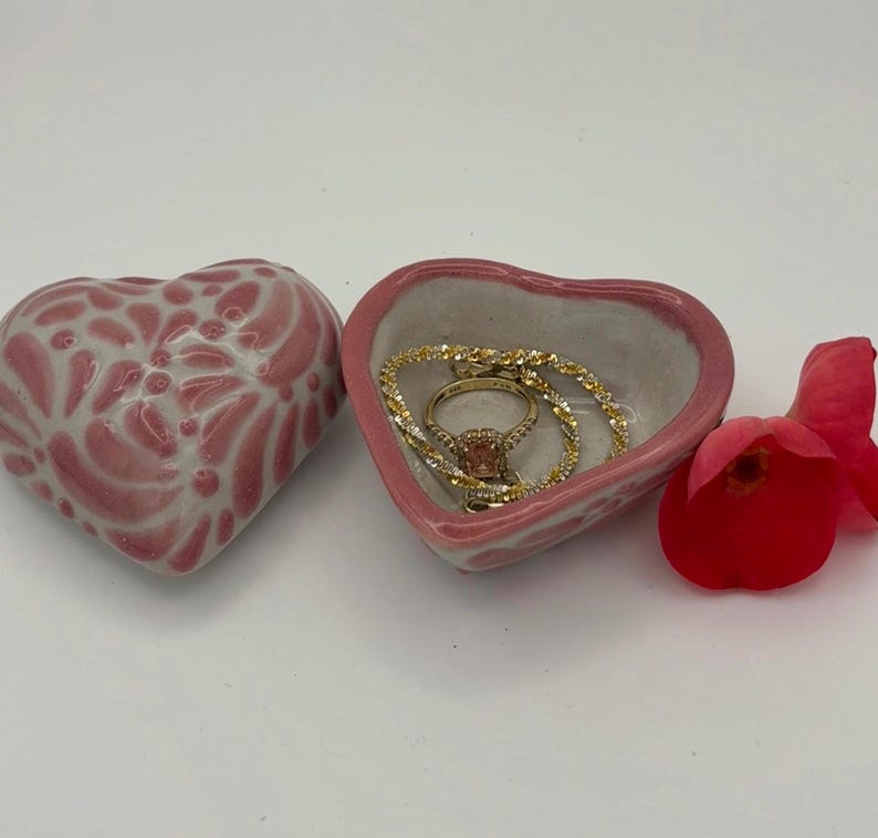Pode incluir: Uma bandeja de cer&acirc;mica rosa em forma de cora&ccedil;&atilde;o com um anel e uma pulseira dourados dentro. Ao lado, uma segunda bandeja em forma de cora&ccedil;&atilde;o com um design floral. &Agrave; direita, uma rosa vermelha.