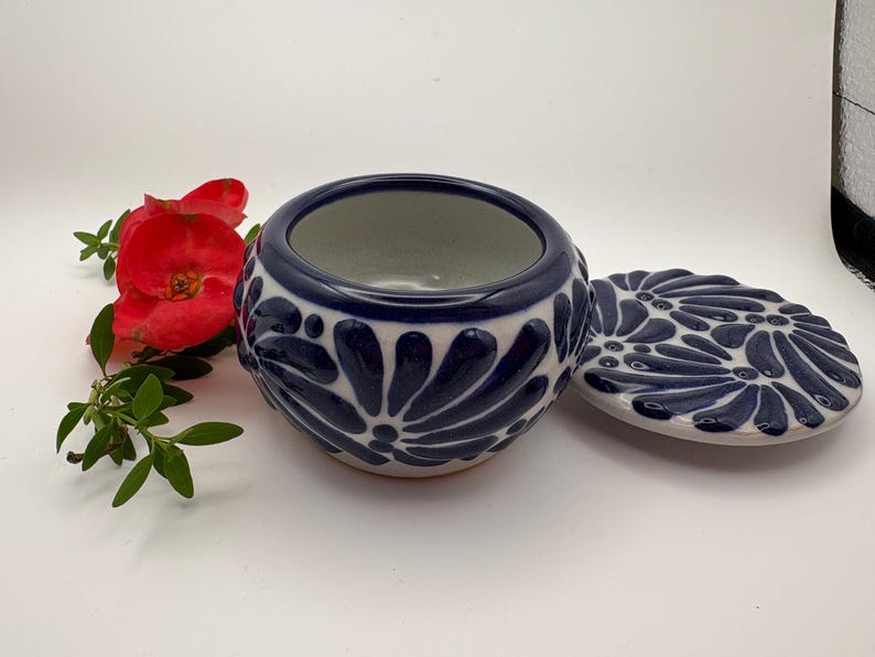 May include: A ceramic jar with a lid, featuring a navy blue and white floral design. The jar is round with a matching lid. A red flower and green leaves are in the background. The jar is likely used for storage or decoration.