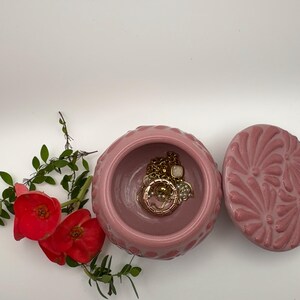 May include: A pink ceramic jewelry box with a lid, containing gold necklaces and bracelets. The box has a textured exterior and a floral design on the lid. Red flowers and green leaves are arranged near the box.