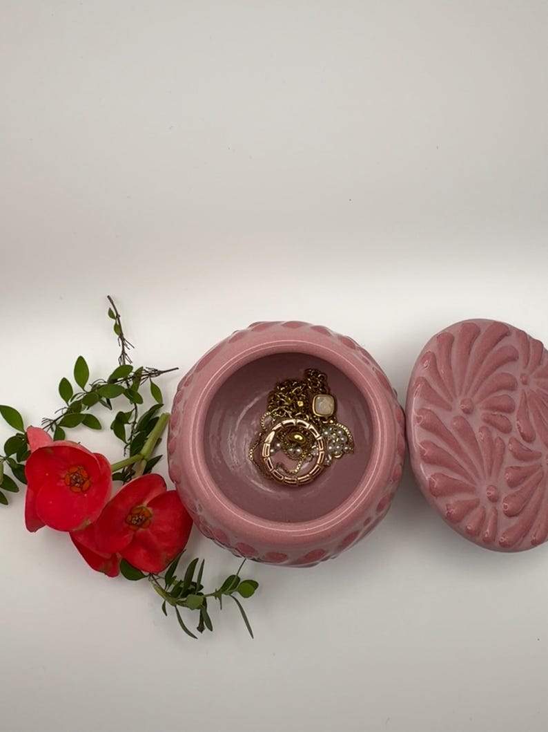 May include: A pink ceramic jewelry box with a lid, containing gold necklaces and bracelets. The box has a textured exterior and a floral design on the lid. Red flowers and green leaves are arranged near the box.