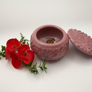 May include: A pink ceramic jewelry box with a lid, featuring a floral design. The box is open, revealing gold jewelry inside. Red flowers and green leaves are arranged beside the box, creating a decorative display.