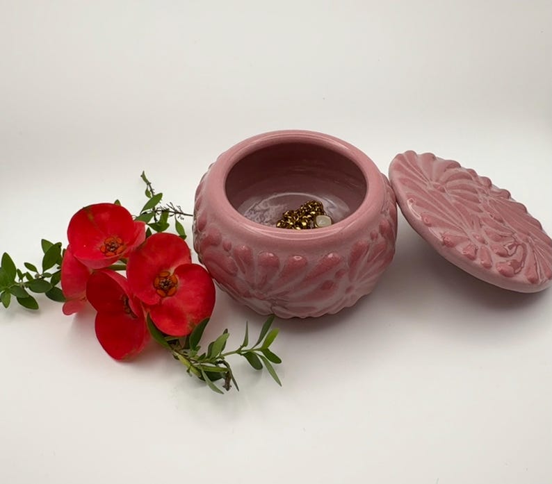 May include: A pink ceramic jewelry box with a lid, featuring a floral design. The box is open, revealing gold jewelry inside. Red flowers and green leaves are arranged beside the box, creating a decorative display.