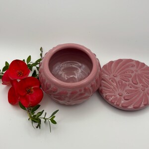 May include: A pink ceramic trinket box with a matching lid. The box and lid feature a raised floral design. Red flowers and green leaves are arranged to the left of the box on a white background.