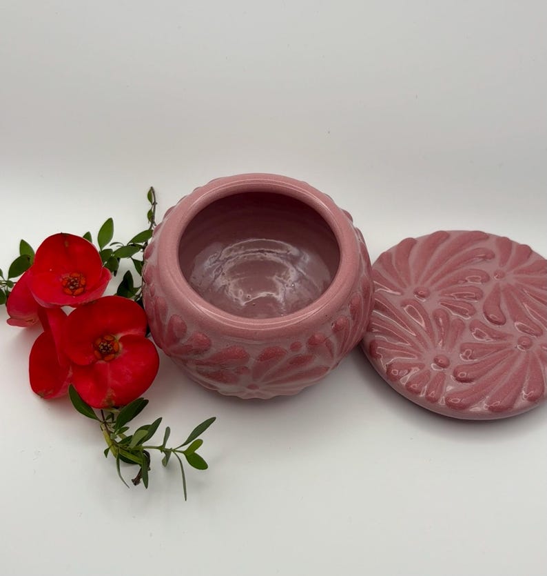 May include: A pink ceramic trinket box with a matching lid. The box and lid feature a raised floral design. Red flowers and green leaves are arranged to the left of the box on a white background.