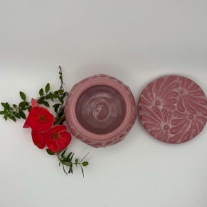 May include: A pink ceramic jar with a matching lid. The jar has a textured pattern around the rim and a smooth interior. The lid features a floral design. Red flowers and green leaves are in the background.