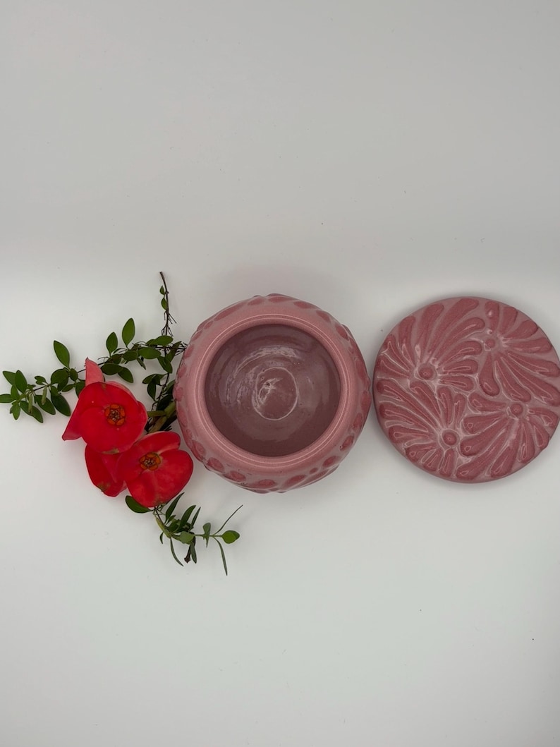 May include: A pink ceramic jar with a matching lid. The jar has a textured pattern around the rim and a smooth interior. The lid features a floral design. Red flowers and green leaves are in the background.
