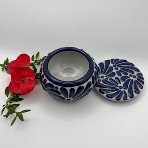 May include: A ceramic bowl with a lid, featuring a navy blue and white floral design. The bowl is round with a textured exterior. The lid is placed next to the bowl. Red flowers and green leaves are in the background.