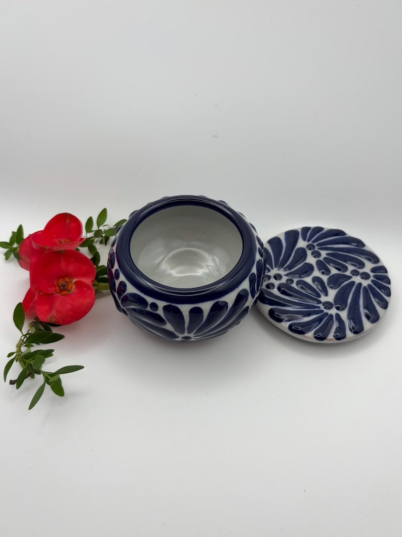 May include: A ceramic bowl with a lid, featuring a navy blue and white floral design. The bowl is round with a textured exterior. The lid is placed next to the bowl. Red flowers and green leaves are in the background.