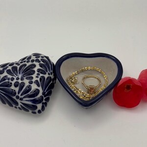 Pode incluir: Uma caixa de bugigangas em forma de cora&ccedil;&atilde;o com um design floral azul e branco, ao lado de um prato em forma de cora&ccedil;&atilde;o aberto com um anel e uma pulseira de ouro. Uma flor vermelha tamb&eacute;m est&aacute; presente.
