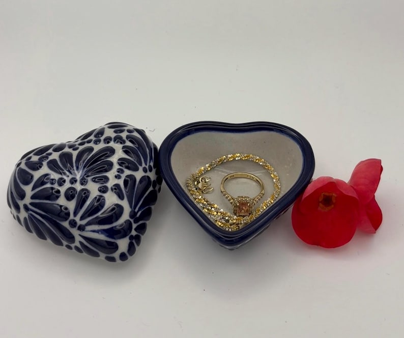 Pode incluir: Uma caixa de bugigangas em forma de cora&ccedil;&atilde;o com um design floral azul e branco, ao lado de um prato em forma de cora&ccedil;&atilde;o aberto com um anel e uma pulseira de ouro. Uma flor vermelha tamb&eacute;m est&aacute; presente.