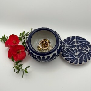 May include: A small, round, ceramic jewelry box with a navy blue and white floral design. The lid is off, revealing gold jewelry inside. Red flowers and greenery are arranged nearby.