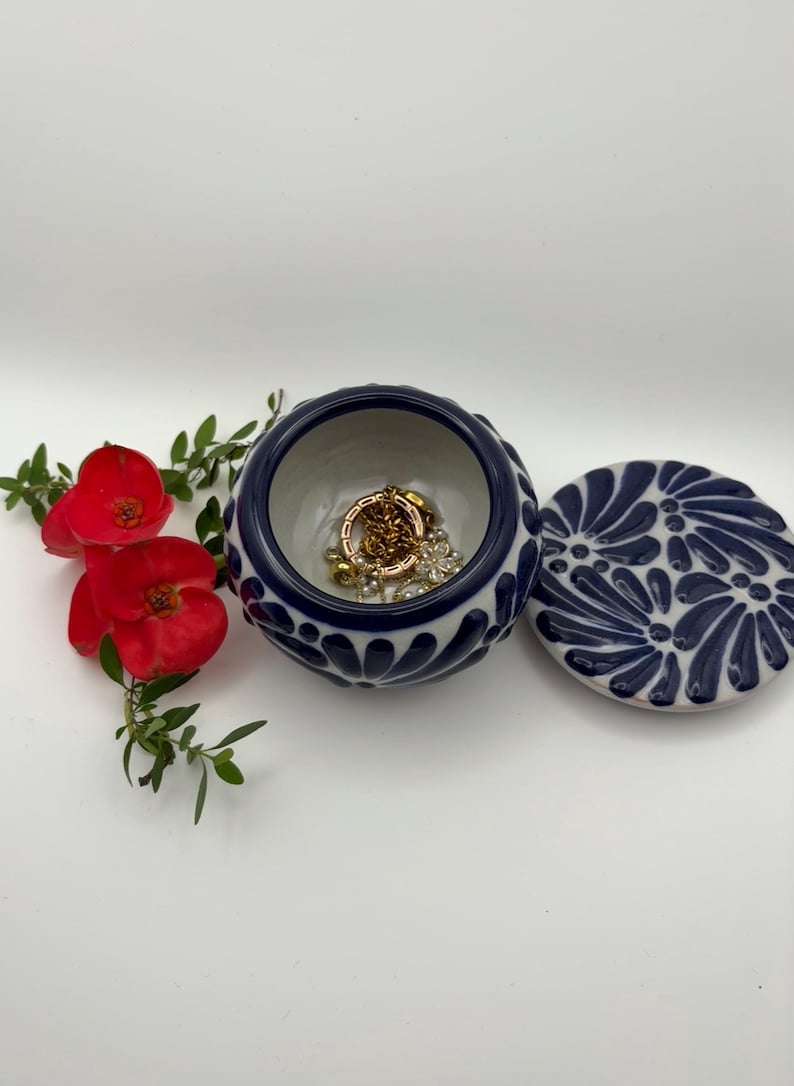 May include: A small, round, ceramic jewelry box with a navy blue and white floral design. The lid is off, revealing gold jewelry inside. Red flowers and greenery are arranged nearby.