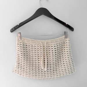 May include: Cream-colored crochet skirt with a drawstring waist and tassel detail, hanging on a black wooden hanger. The skirt has a diamond-shaped open weave design and a scalloped hemline. A casual, beach-inspired garment.