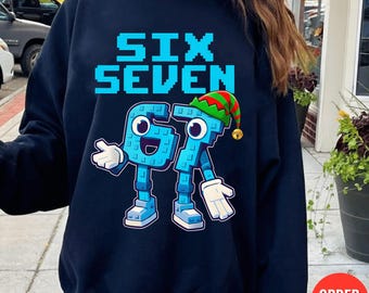 Puede incluir: Sudadera azul marino con las palabras "SIX SEVEN" en fuente pixelada azul claro. Debajo, los números 6 y 7 se representan como personajes de dibujos animados, con brazos, piernas y un gorro de elfo navideño en el número 7.