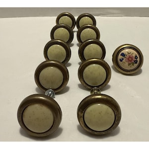 May include: A set of nine round cabinet knobs with a metallic brass-toned finish and speckled cream-colored centers. One knob features a floral design with pink and blue flowers on a white background, while the others have plain cream centers.