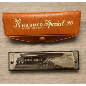 Puede incluir: Una armónica Hohner Special 20 con una placa de cubierta de metal plateado que presenta el texto Hohner Special 20 Marine Band y una C, acompañada de un estuche de piel sintética texturizada de color tostado con el logo y texto Hohner en dorado.