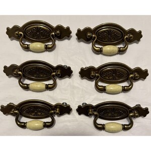 May include: Six antique bronze drawer pulls with cream-colored ceramic handles. Each pull features an oval design with a floral pattern and decorative edges. The pulls are arranged in two rows of three.