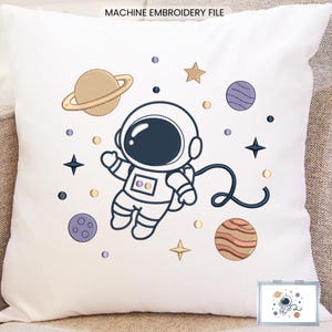 May include: A white square throw pillow featuring a machine embroidery design of an astronaut floating in space. The design includes a tan planet with rings, a purple planet, a striped orange planet, gold stars, and small dots. Text reads MACHINE EMBROIDERY FILE.
