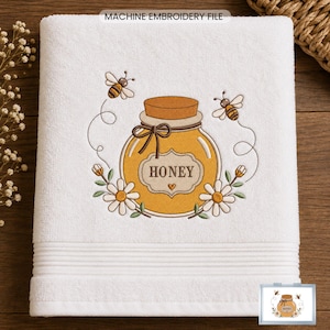 Op de afbeelding: Een witte handdoek met een machinaal borduursel van een honingpot met een kurken deksel, twee bijen en witte madeliefjes. De pot heeft een etiket met de tekst HONEY en een klein hartje. De details zijn uitgevoerd in bruin stiksel.