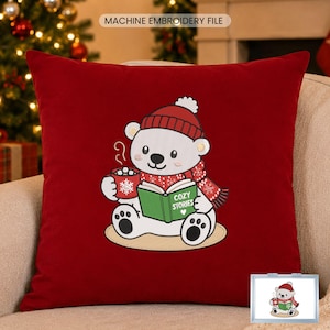 May include: A deep red square throw pillow featuring an embroidered white polar bear wearing a red beanie and scarf. The bear holds a red mug with a snowflake and a green book with the text COZY STORIES. A small inset shows the same design.