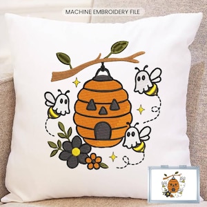 May include: A white throw pillow featuring a machine embroidery design of an orange beehive shaped like a jack-o'-lantern, surrounded by three bees wearing white ghost costumes, a brown branch, and black and orange flowers with yellow stars.