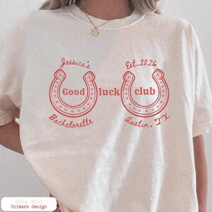 May include: Ivory-colored t-shirt with red text and horseshoe graphics. The shirt features the text "Jessica's Good Luck Bachelorette" and "Est. 2026 Good Luck Club Austin, TX."