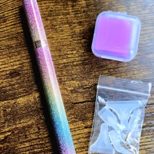May include: A multi-colored pen with a glittery finish, a purple square case, and a small bag of clear accessories are displayed on a wooden surface. The pen has a rainbow gradient design and a gold-colored metal tip.