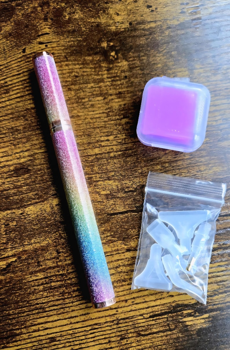 May include: A multi-colored pen with a glittery finish, a purple square case, and a small bag of clear accessories are displayed on a wooden surface. The pen has a rainbow gradient design and a gold-colored metal tip.