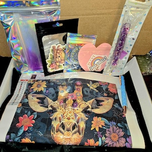 May include: A diamond painting kit featuring a giraffe with floral accents. The kit includes a canvas, various colored gems, a pen, and other accessories. The image also shows a heart-shaped sticker that says "Just smile!"