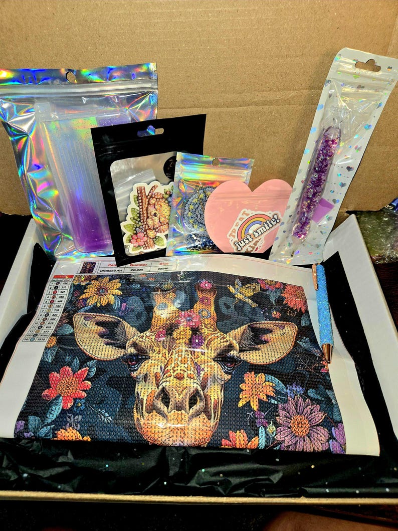 May include: A diamond painting kit featuring a giraffe with floral accents. The kit includes a canvas, various colored gems, a pen, and other accessories. The image also shows a heart-shaped sticker that says "Just smile!"
