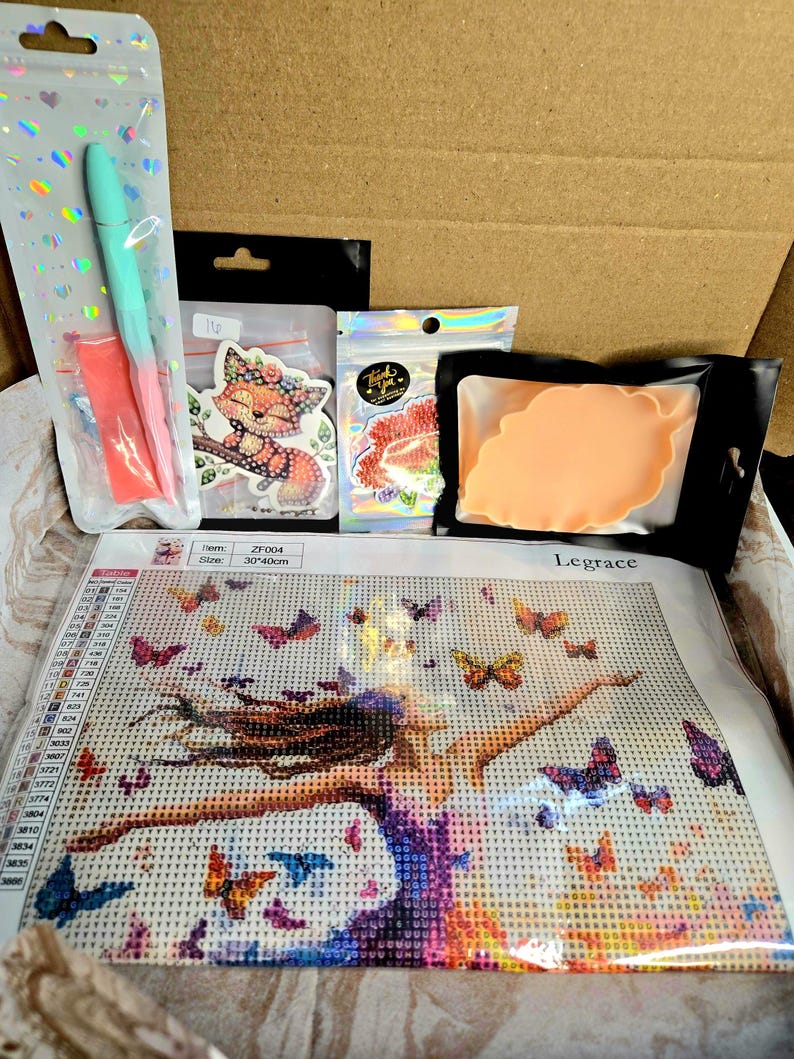 May include: An open cardboard box containing a diamond painting kit with a woman and butterflies design, along with a pen, stickers, and a resin mold. The painting is 30 x 40cm. The kit includes various accessories for crafting.