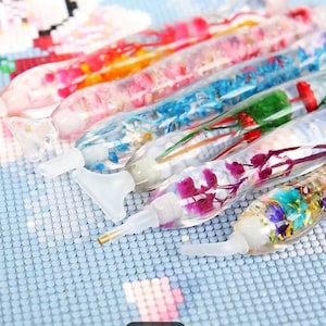 May include: A collection of colorful diamond painting pens filled with dried flowers and glitter. The pens have clear barrels and white tips, and are arranged on a light blue surface. The pens are designed for crafting and art projects.