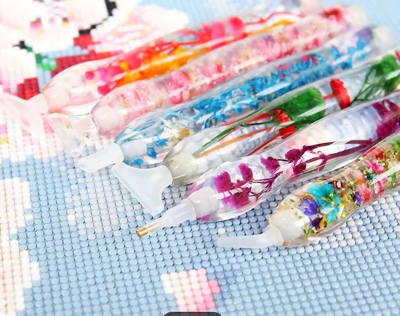 May include: A collection of colorful diamond painting pens filled with dried flowers and glitter. The pens have clear barrels and white tips, and are arranged on a light blue surface. The pens are designed for crafting and art projects.