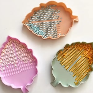 May include: Three leaf-shaped trays in peach, pink, and green, each filled with small, colorful beads. The peach tray holds silver and blue beads, the pink tray has pink and white beads, and the green tray contains yellow beads. The trays are designed for crafting.