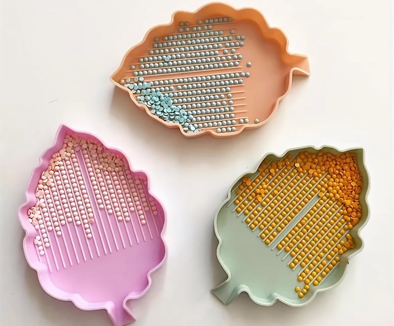 May include: Three leaf-shaped trays in peach, pink, and green, each filled with small, colorful beads. The peach tray holds silver and blue beads, the pink tray has pink and white beads, and the green tray contains yellow beads. The trays are designed for crafting.