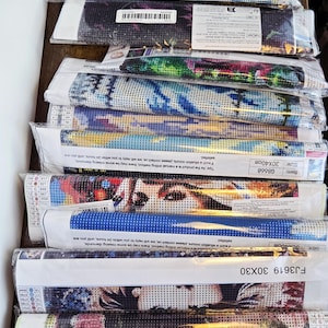 May include: A stack of rolled-up diamond painting kits, each wrapped in clear plastic. The kits feature various colorful designs, including portraits and abstract patterns. The sizes are 30x30 cm and 30x40 cm.