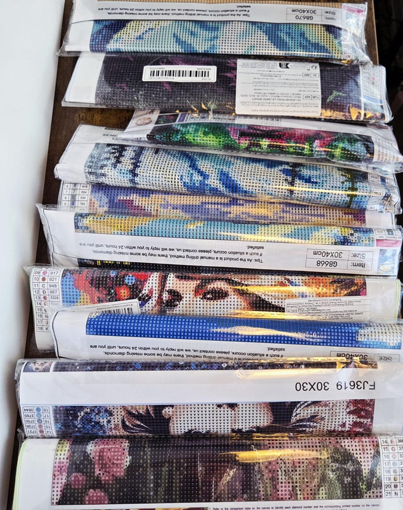 May include: A stack of rolled-up diamond painting kits, each wrapped in clear plastic. The kits feature various colorful designs, including portraits and abstract patterns. The sizes are 30x30 cm and 30x40 cm.