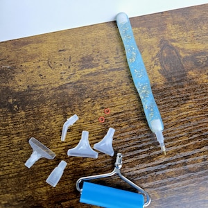 May include: A collection of diamond painting tools, including a blue pen with gold flecks, various clear plastic applicator tips, small red rings, and a blue roller. The items are arranged on a brown wooden surface.