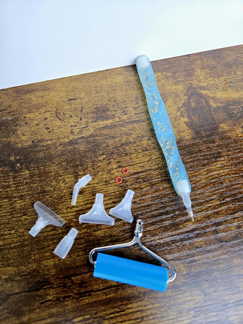 May include: A collection of diamond painting tools, including a blue pen with gold flecks, various clear plastic applicator tips, small red rings, and a blue roller. The items are arranged on a brown wooden surface.