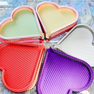 May include: Five heart-shaped trays in various colors: red, purple, silver, yellow, and green. Each tray has a diagonal striped pattern and a braided rope-like border. The trays are arranged in a circular pattern.