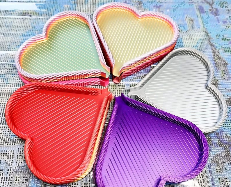 May include: Five heart-shaped trays in various colors: red, purple, silver, yellow, and green. Each tray has a diagonal striped pattern and a braided rope-like border. The trays are arranged in a circular pattern.