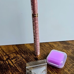 May include: A pink and gold pen with a textured pattern, a small purple square container, and a small bag of white accessories are displayed on a wooden surface. The pen has a metallic finish.