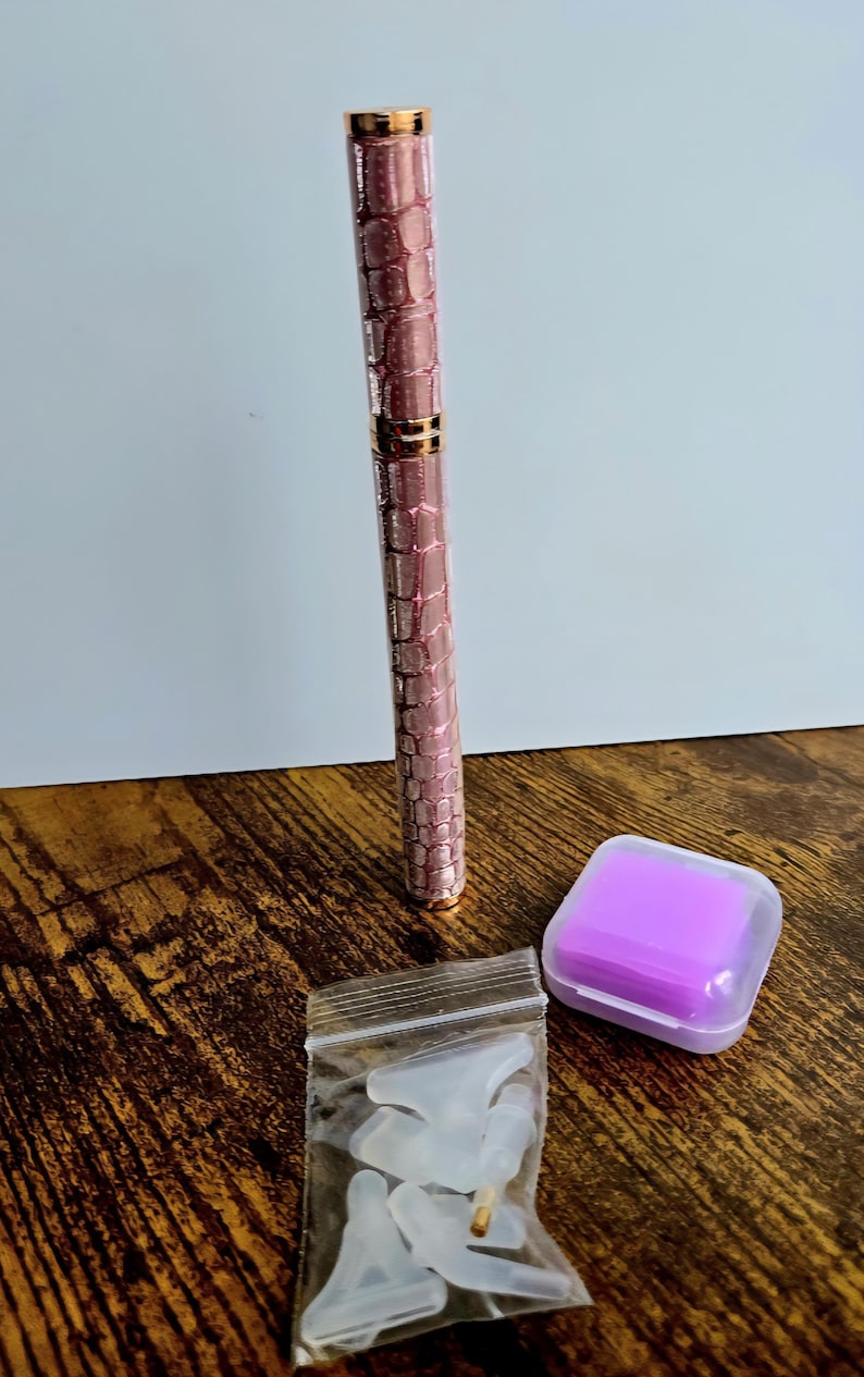 May include: A pink and gold pen with a textured pattern, a small purple square container, and a small bag of white accessories are displayed on a wooden surface. The pen has a metallic finish.